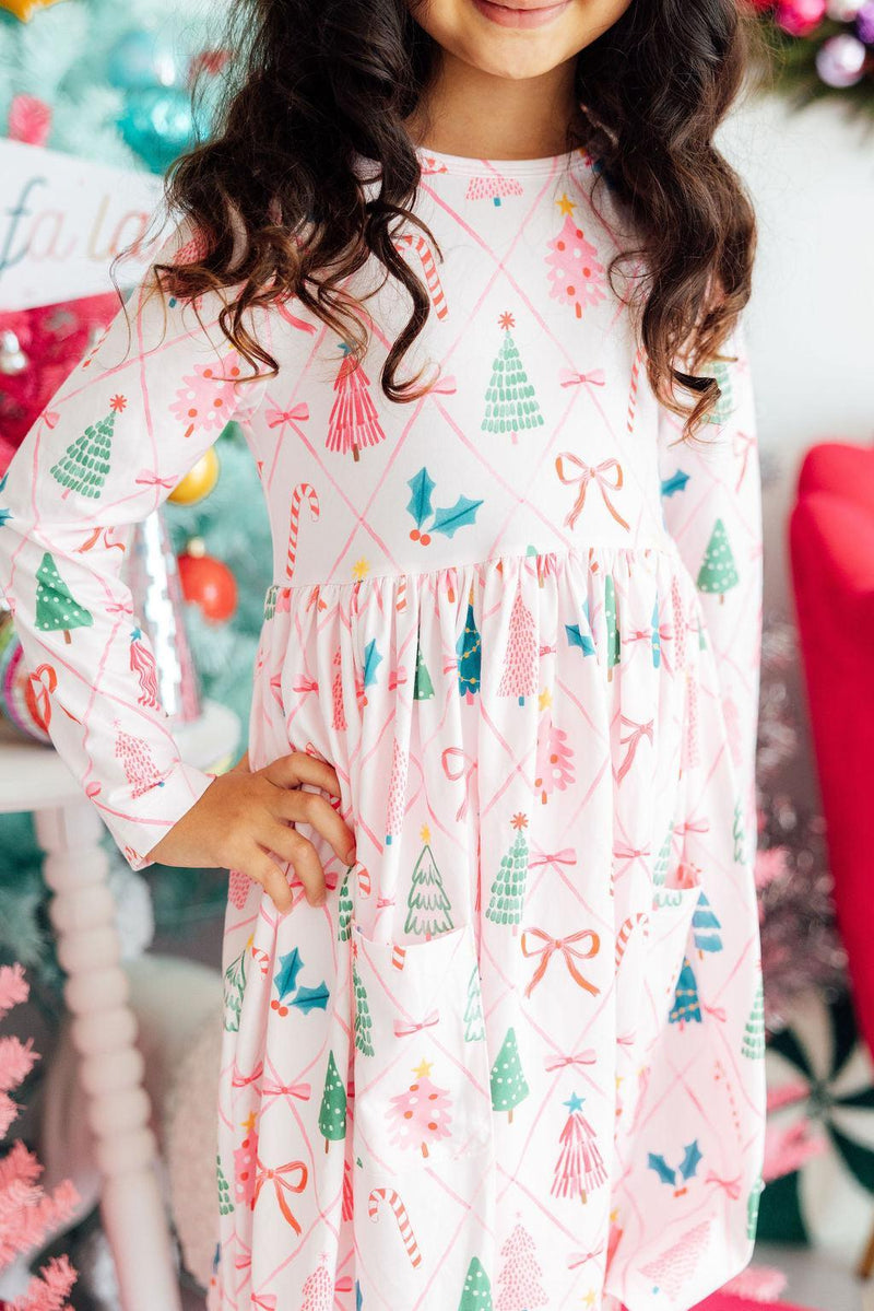 Girls' long sleeve pocket twirl dress featuring festive trees, candy canes, and bows on a white background with pink diamond grid.