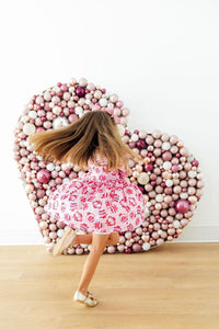 Girl twirling in a pink long-sleeve pocket twirl dress beside a heart-shaped wall of pink ornaments.