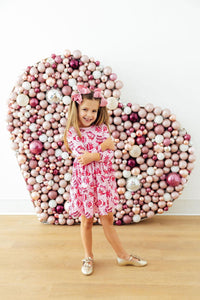 Girl in Vintage Cakes Long Sleeve Pocket Twirl Dress, pink, full-circle twirl skirt, posing by ornament backdrop.