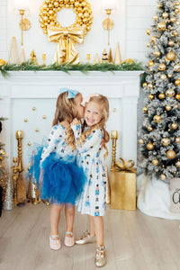 Girls in long sleeve nutcracker print dresses with blue tulle skirts celebrating near a gold holiday wreath and Christmas tree.