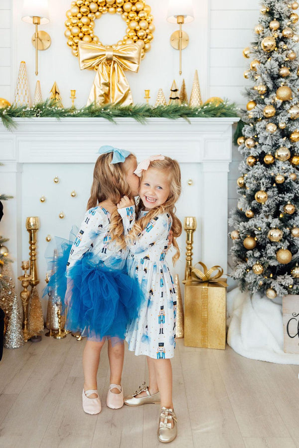 Girls in long sleeve nutcracker print dresses with blue tulle skirts celebrating near a gold holiday wreath and Christmas tree.