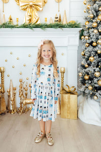 Girl in long sleeve nutcracker print twirl dress with pockets standing near gold Christmas tree and decorated fireplace.