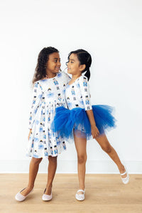 Two girls smile while wearing long sleeve nutcracker print twirl dresses; one features a full skirt and the other a blue tulle petticoat.