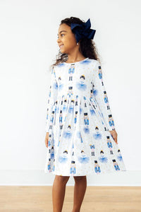 Girl in white long sleeve twirl dress with blue ballerina and nutcracker print, full circle skirt, and navy bow headband.