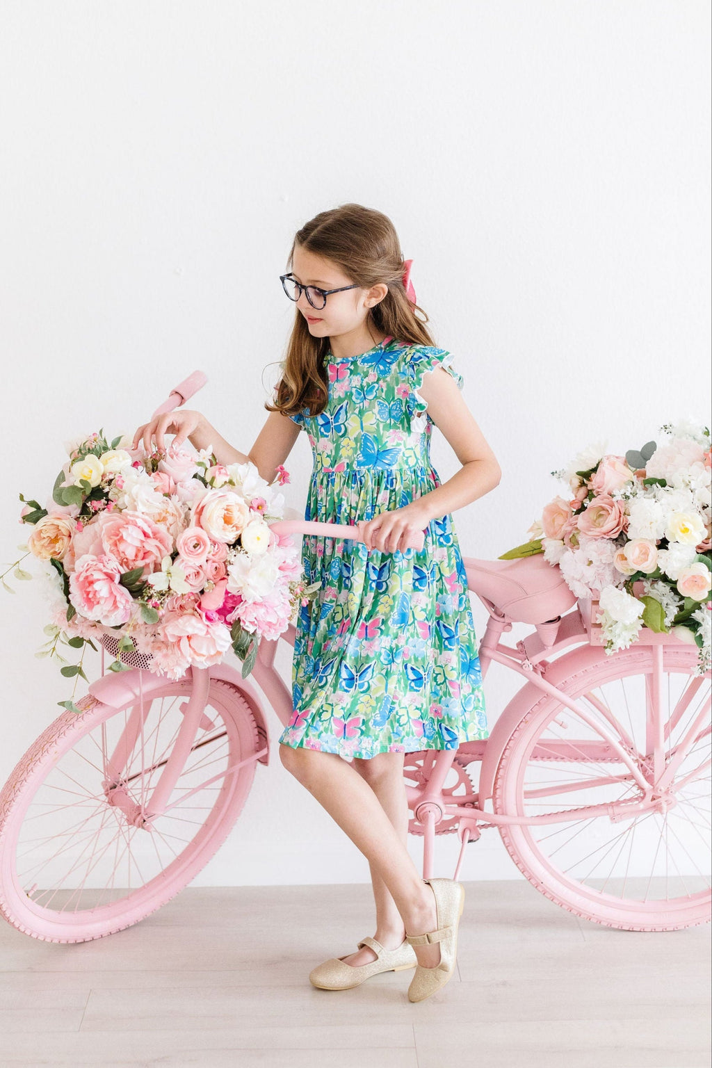 A young girl wearing the Watercolor Butterflies Flutter Sleeve Twirl Dress poses next to a pink bicycle adorned with flowers, showcasing the vibrant butterfly print and flutter sleeves.