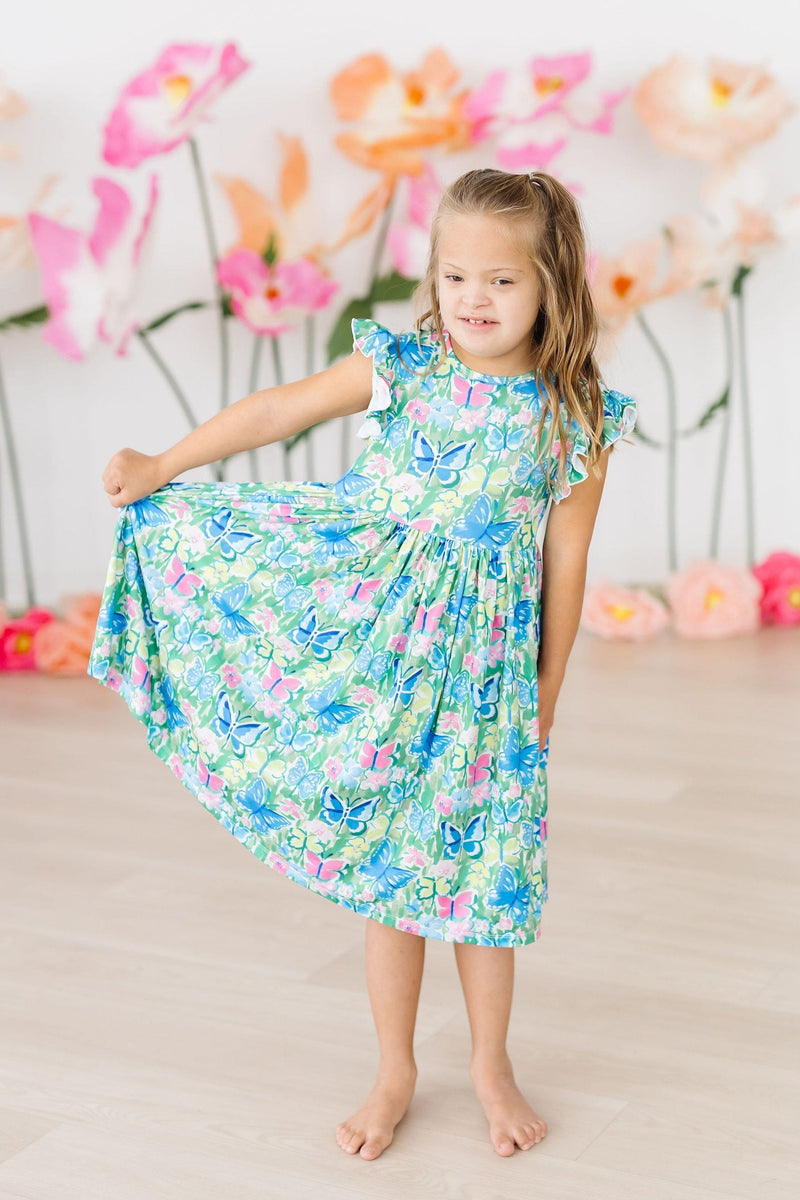 A young girl twirls in the Watercolor Butterflies Flutter Sleeve Twirl Dress, displaying the dress's flowy silhouette and butterfly print, surrounded by colorful floral decorations.