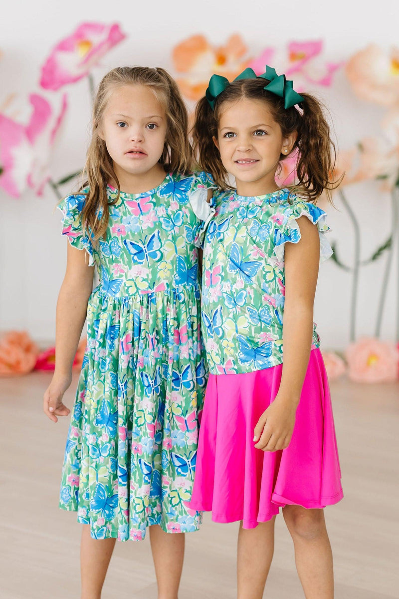 Two girls smile while wearing the Watercolor Butterflies Flutter Sleeve Twirl Dress, with one in a matching dress and the other in a pink skirt, highlighting the playful design and colorful butterfly pattern.