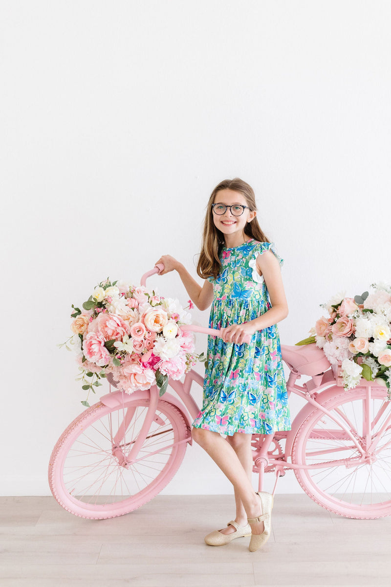 Girl posing with a pink bicycle adorned with flowers, wearing the Watercolor Butterflies Flutter Sleeve Twirl Dress, radiating style and charm.
