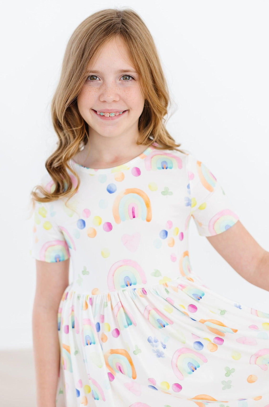 Girl wearing the Watercolor Rainbows S/S Pocket Twirl Dress from Mila & Rose, showcasing a vibrant rainbow print and smiling in a bright, cheerful pose.