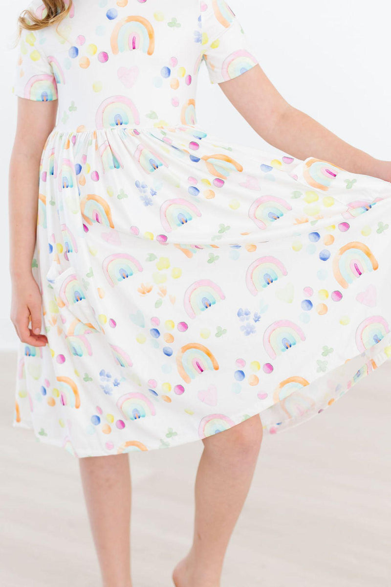 Close-up of the Watercolor Rainbows S/S Pocket Twirl Dress from Mila & Rose, featuring the playful rainbow and dot patterns as the girl holds the skirt.