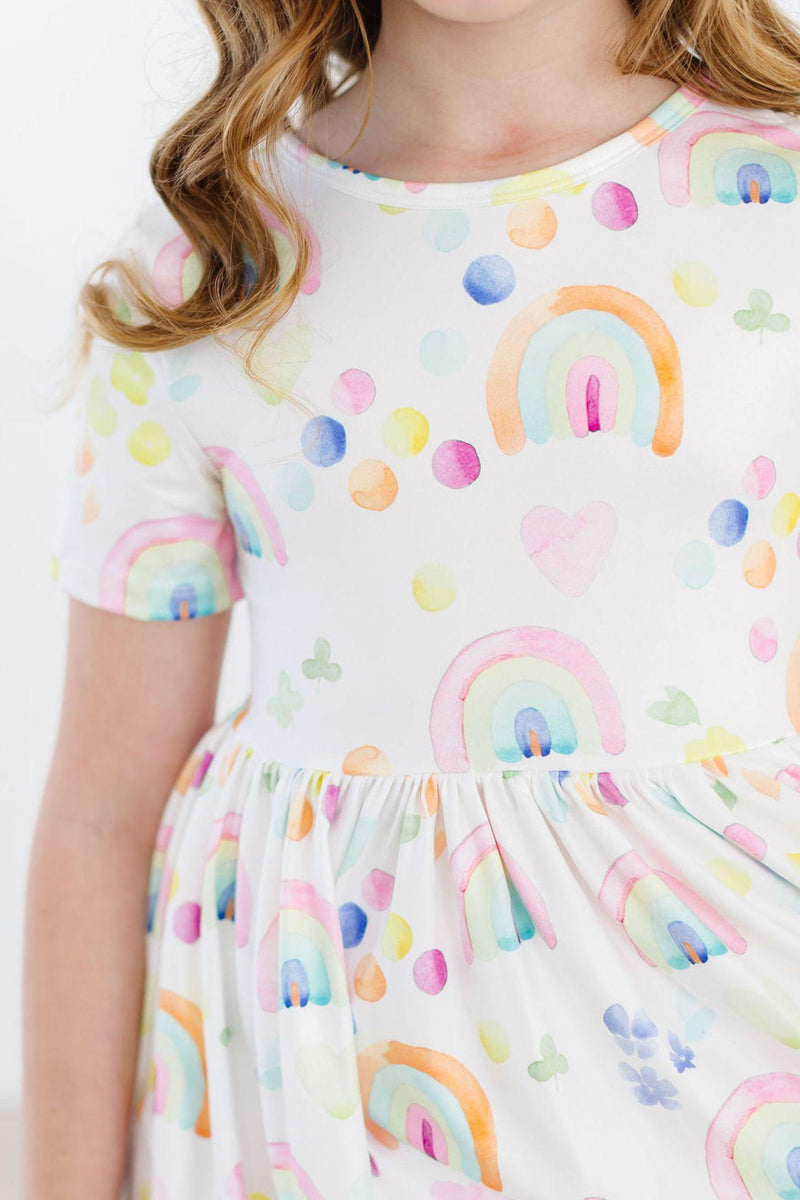 Close-up of the Watercolor Rainbows S/S Pocket Twirl Dress, featuring a colorful rainbow print on a soft fabric, worn by a girl with wavy hair.