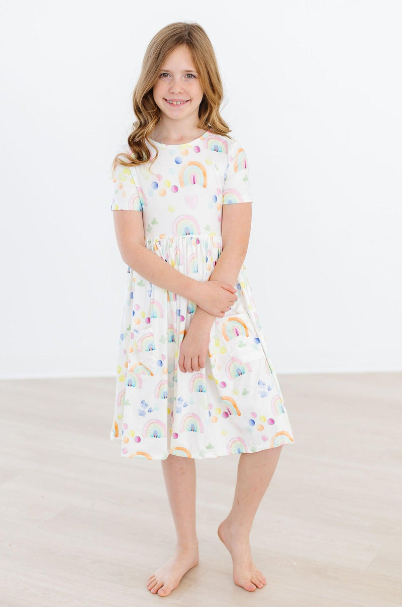 Girl wearing the Watercolor Rainbows S/S Pocket Twirl Dress, striking a pose with her hands crossed, showcasing the vibrant print and twirl skirt.