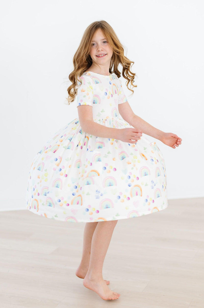 Girl joyfully posing in the Watercolor Rainbows S/S Pocket Twirl Dress, emphasizing the lively rainbow patterns and twirly design.