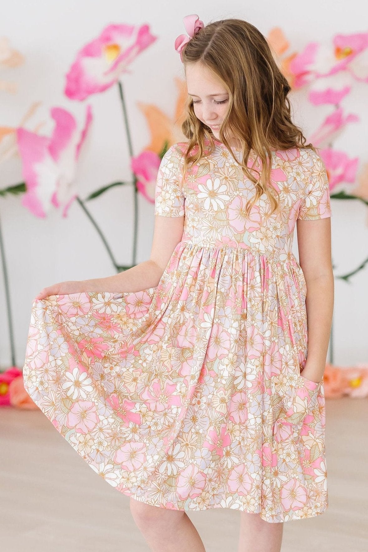 Girl twirling a pink and white floral short sleeve pocket dress with a gathered skirt against a floral backdrop.