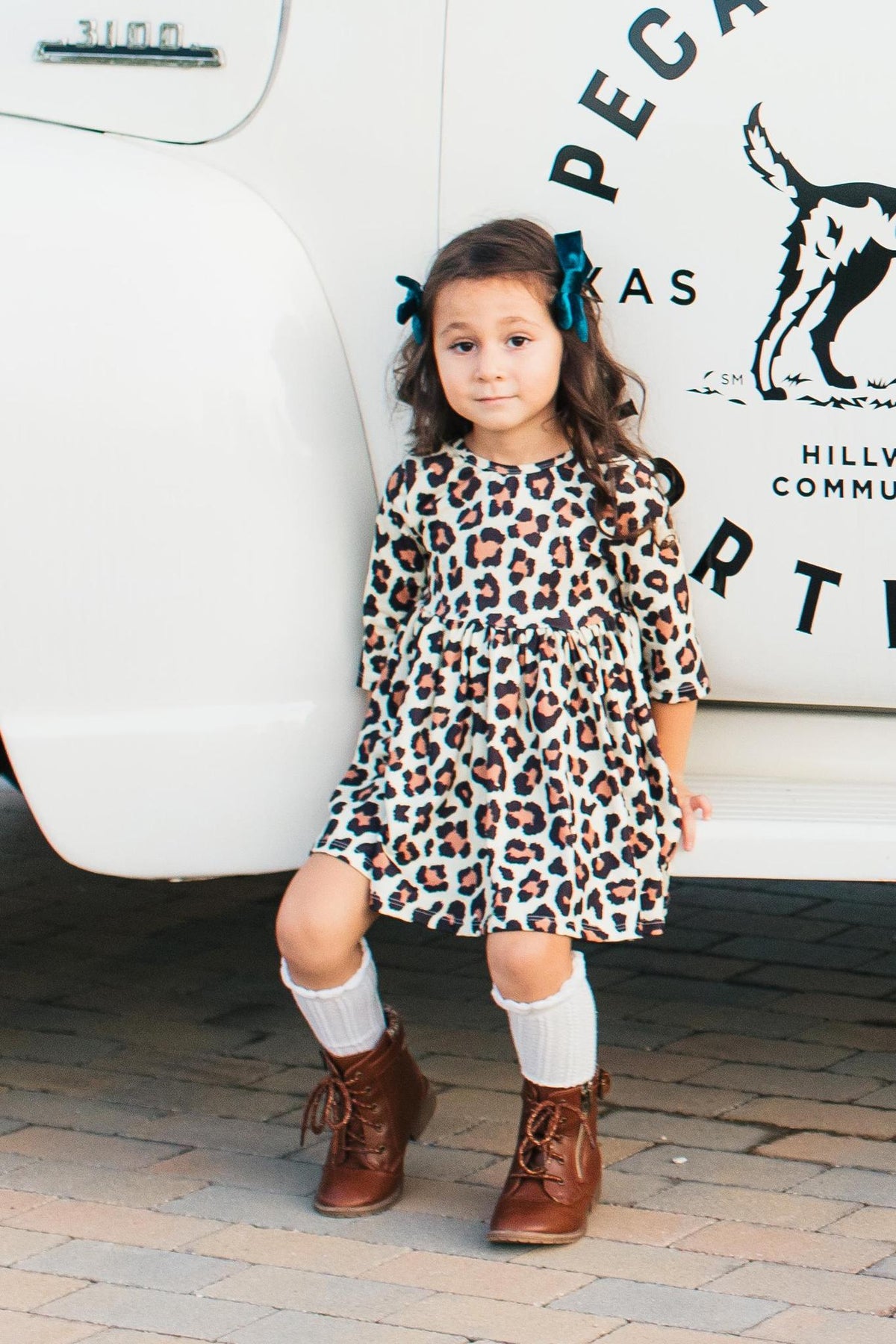 Toddler girl wearing a leopard print 3/4 sleeve twirl dress with pockets, styled with brown boots and leg warmers.