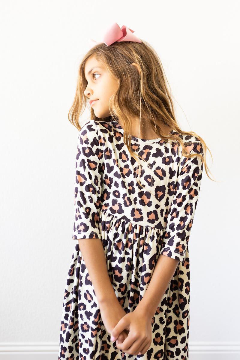Child wearing a leopard print 3/4 sleeve twirl dress featuring a scoop back and side pocket, paired with a pink hair bow.