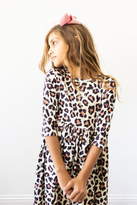 Child wearing a leopard print 3/4 sleeve twirl dress featuring a scoop back and side pocket, paired with a pink hair bow.