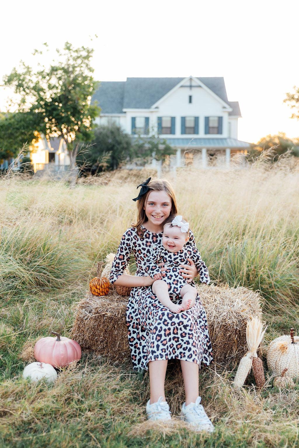 Girl in leopard print 3/4 sleeve pocket twirl dress holding baby in matching outfit on hay bale with pumpkins.