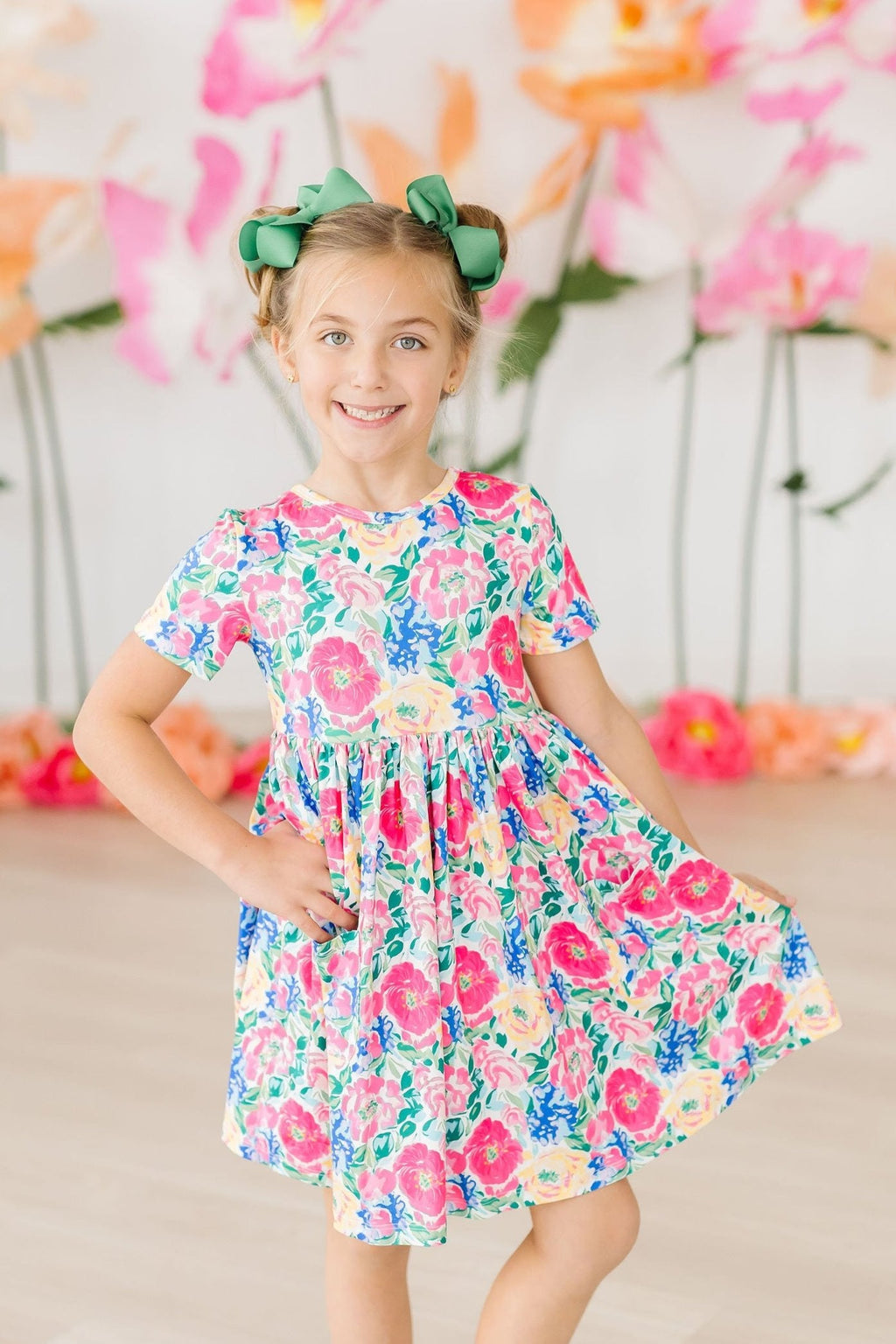 Wildflower Wishes S/S Pocket Twirl Dress on a smiling girl posing against a colorful flower backdrop, showcasing the vibrant floral design and twirl factor.
