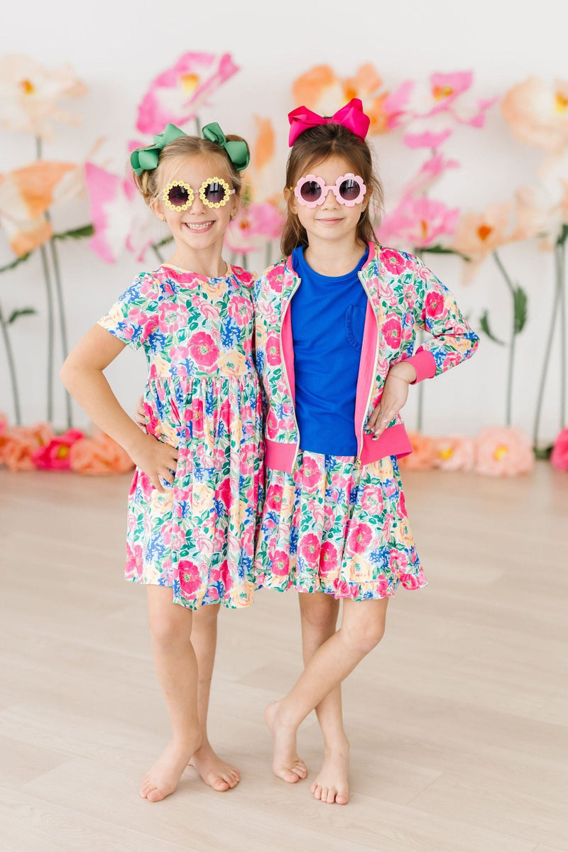 Two girls wearing the Wildflower Wishes S/S Pocket Twirl Dress with sunglasses and playful smiles, highlighting the dress's colorful design and style versatility.