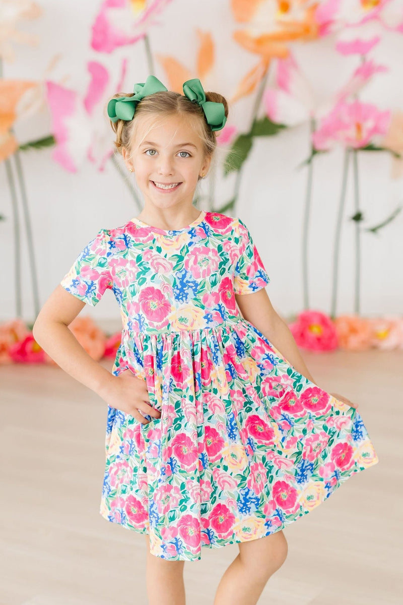Wildflower Wishes S/S Pocket Twirl Dress on a smiling girl posing against a colorful flower backdrop, showcasing the vibrant floral design and twirl factor.
