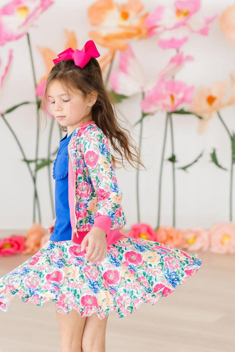 Young girl twirling in a vibrant floral Wildflower Wishes Satin Jacket over a blue top and matching floral skirt, against a backdrop of pink flowers, showcasing a playful and stylish look.