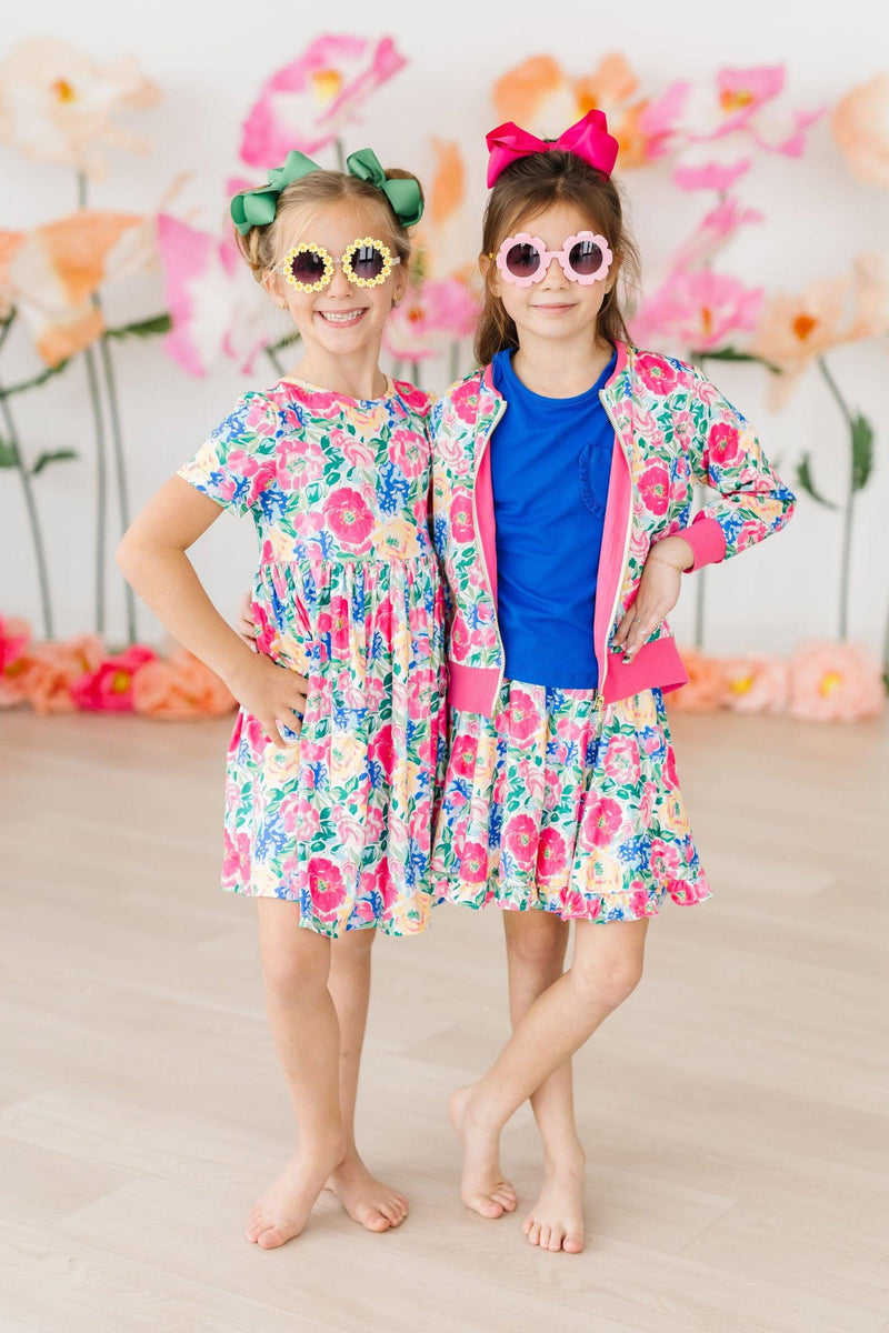 Two girls modeling the Wildflower Wishes Satin Jacket and coordinating floral dresses, smiling confidently with fun accessories and sunglasses, in a colorful floral-themed setting.
