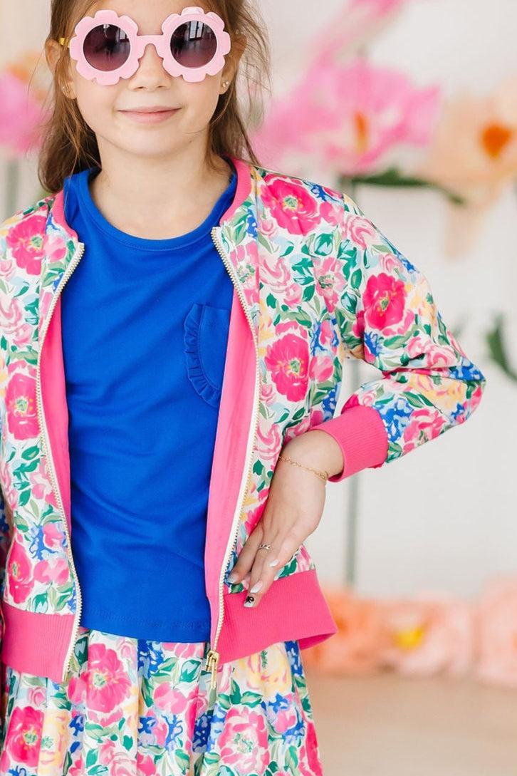 A young girl modeling the Wildflower Wishes Satin Jacket from Mila & Rose, paired with a blue top and colorful skirt. She is wearing stylish pink sunglasses, showcasing a trendy look.