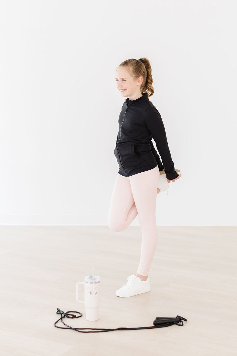 A girl in Zen Pink Active Legging by Mila & Rose, stretching while holding onto her foot, wearing a black jacket, set against a minimalistic white background.