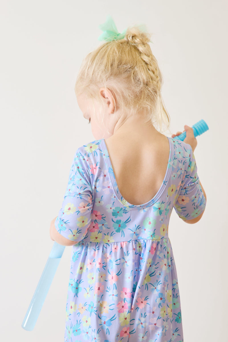 The back of a toddler wearing the Petal Party Wide Leg Jumpsuit by Mila & Rose, featuring a playful floral pattern and a scoop back. She is holding a blue toy, emphasizing a carefree moment.