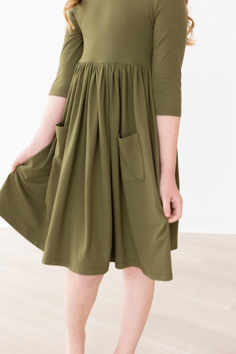 Close-up of the Olive 3/4 Sleeve Pocket Twirl Dress by Mila & Rose, featuring a little girl holding the dress and displaying the pockets and fabric drape.