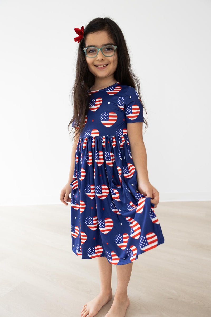 A smiling girl in the 'I Heart the USA' Short Sleeve Pocket Twirl Dress for Girls, displaying the beautiful heart patterns with American flags, perfect for fun and playful twirls.