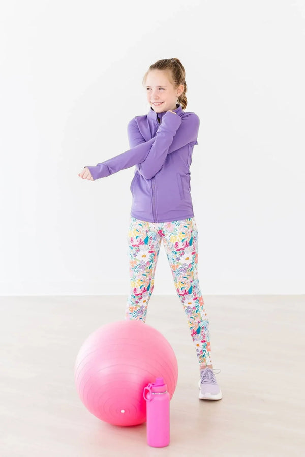 Girls colorful floral active leggings from Mila and Rose paired with purple jacket stretching indoors