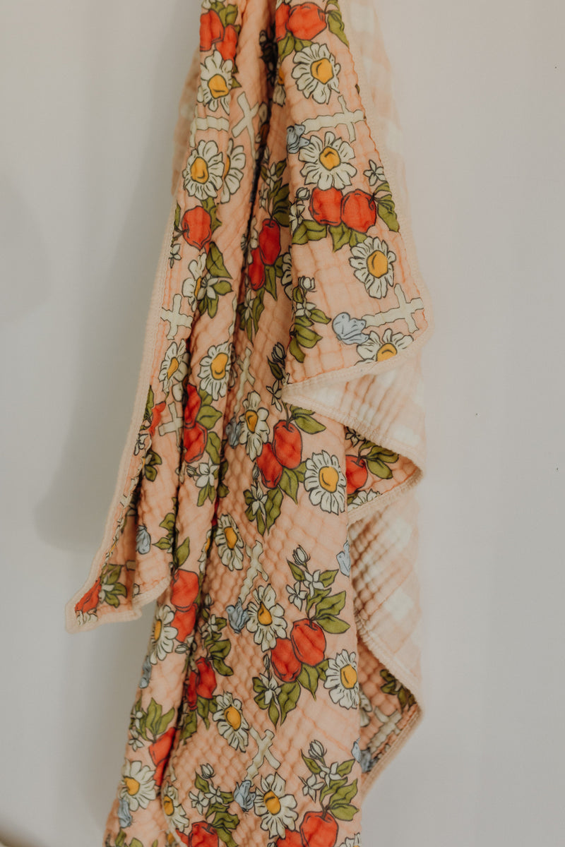 Muslin swaddle draped against white wall showing lightweight fabric and floral cross pattern