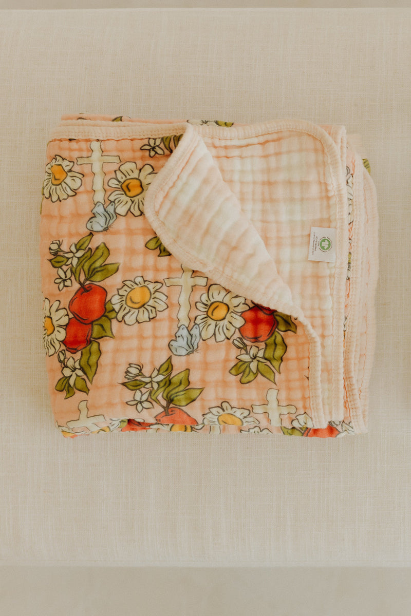 Folded organic cotton baby blanket with peach gingham, white daisies, red apples, and crosses