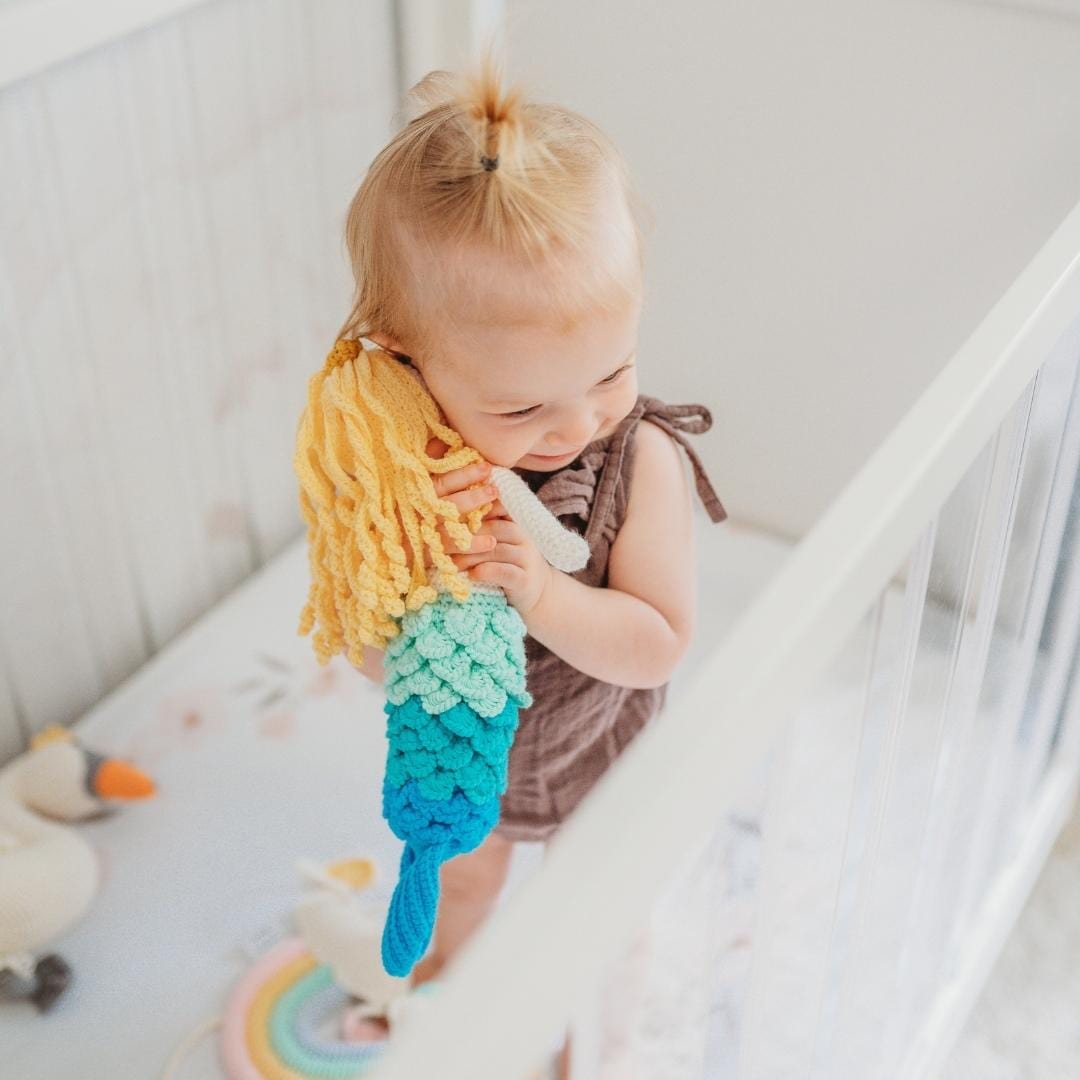 Toddler holding soft crochet mermaid doll with blonde curls and blue-green tail in white crib.