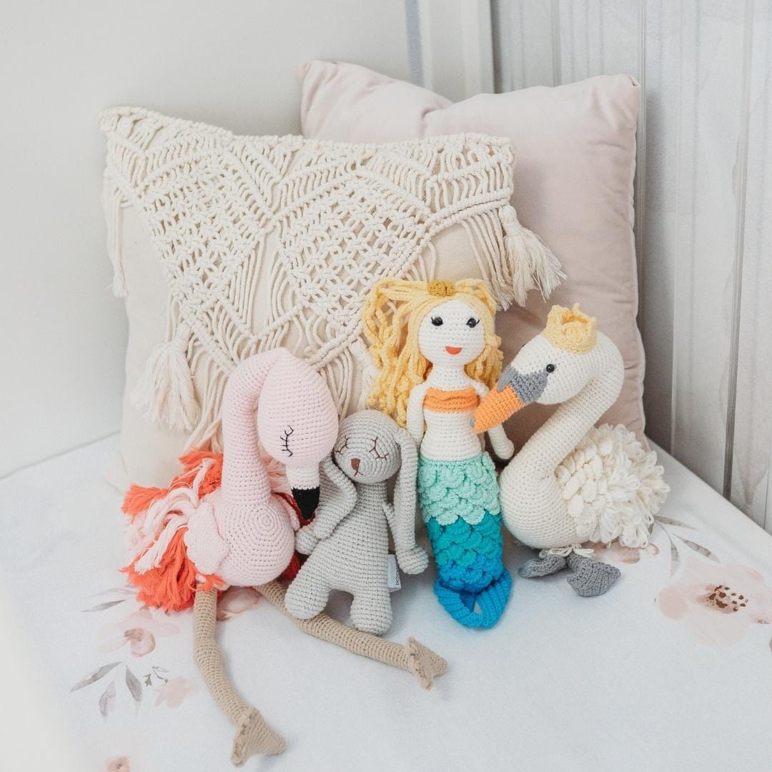 Blonde curly-haired mermaid doll with blue-green tail and tiny crown sitting among crocheted flamingo, bunny, and swan toys.