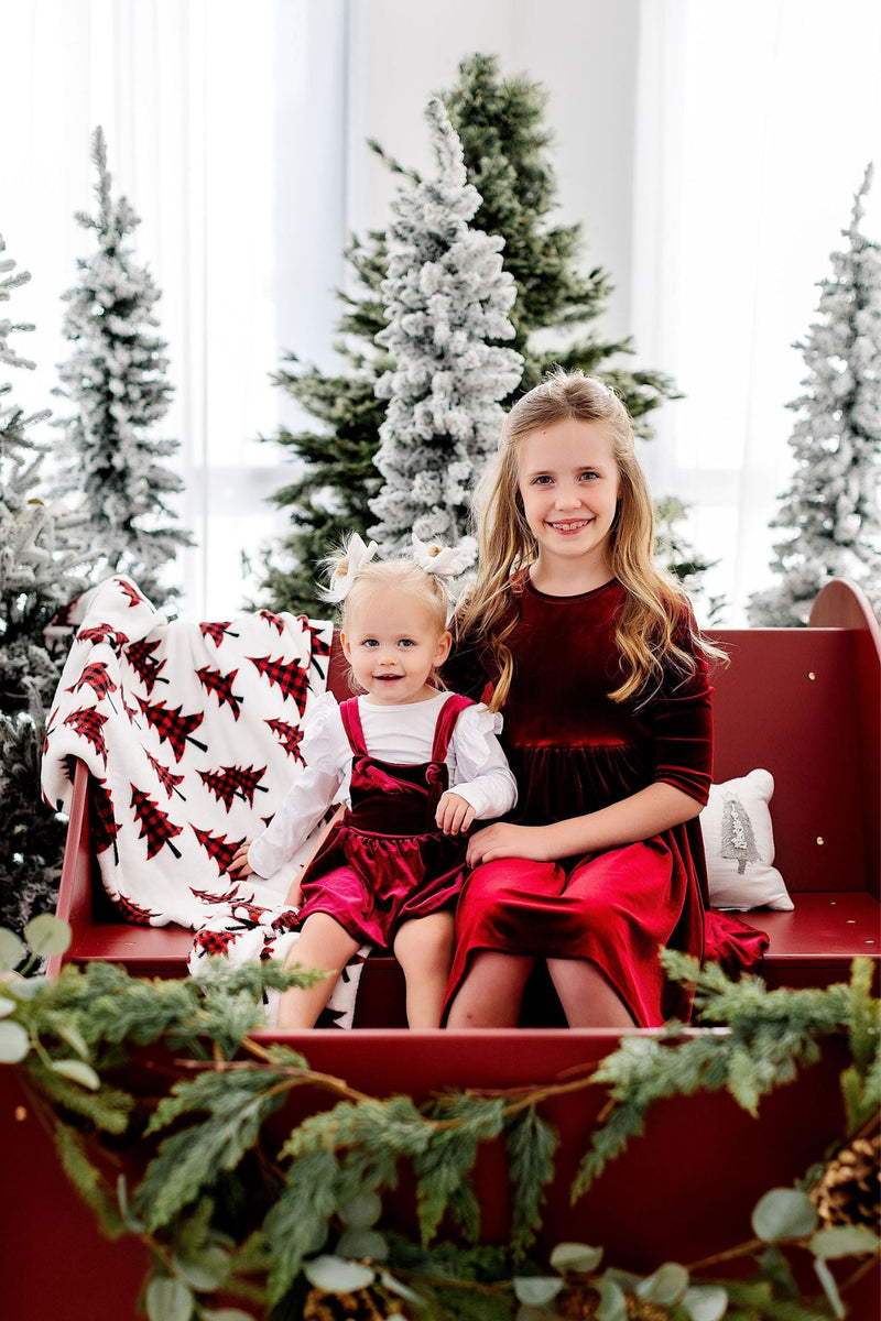 Two sisters happily posing in festive Cranberry Velvet Twirl Dresses from Mila & Rose®, surrounded by snowy trees and a cozy red sleigh, perfect for holiday memories.