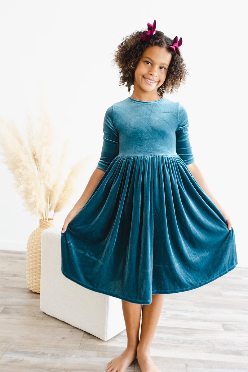 Girl wearing the Denim Blue Velvet Twirl Dress for Girls, smiling and posing while holding the skirt, emphasizing the twirl-friendly design.