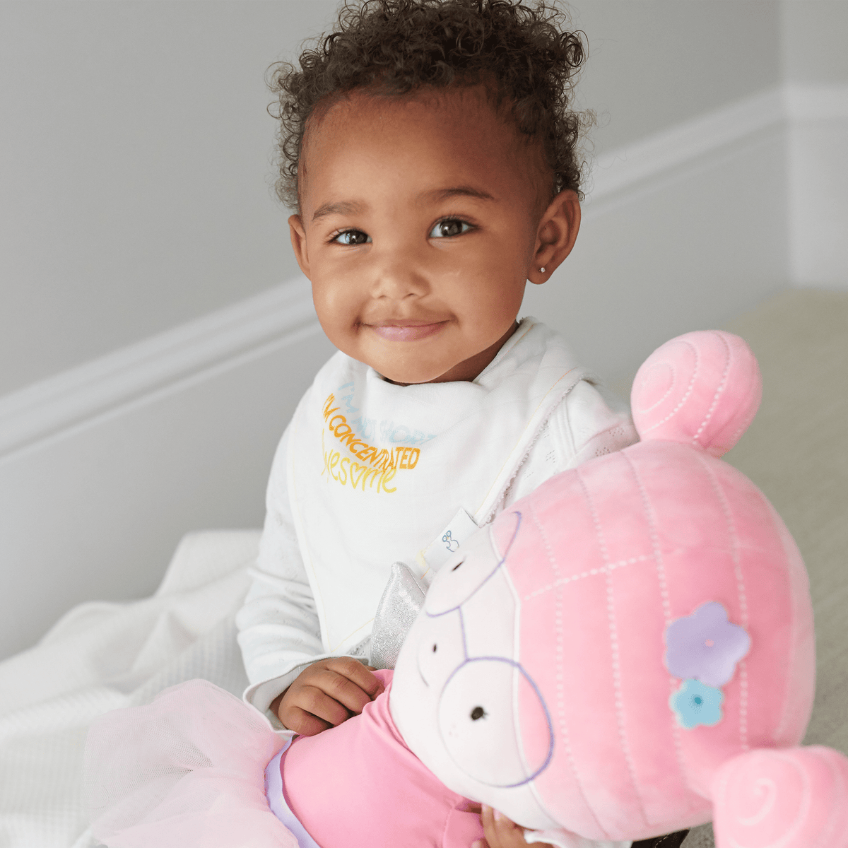 toddler wearing white baby bibs with yellow text holding pink plush toy with flower details
