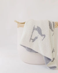 Creamy white baby blanket with charcoal heron print draped over a woven basket, showcasing coastal elegance and soft texture.