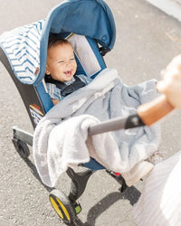 Smiling baby in a blue stroller wrapped in a soft, light charcoal and white heron print luxury receiving blanket.