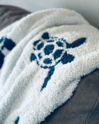 Soft white baby blanket featuring a navy blue sea turtle design, capturing the serene allure of coastal waves and sandy shores.
