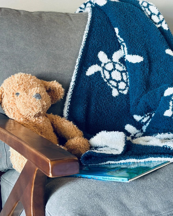 Soft navy baby blanket with white sea turtles draped on a grey chair beside a teddy bear and book.