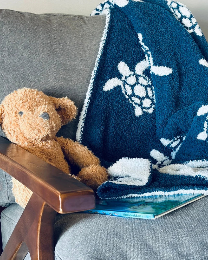 Soft navy baby blanket with white sea turtles draped on a grey chair beside a teddy bear and book.
