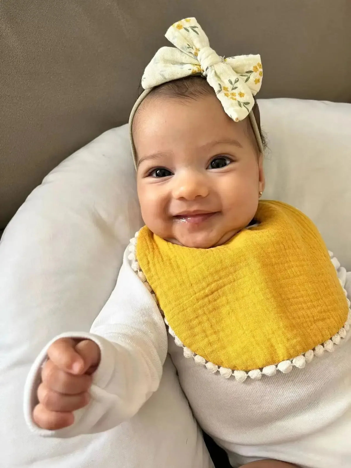 smiling baby wearing yellow baby flower bib and headband set with floral bow lying on white cushion