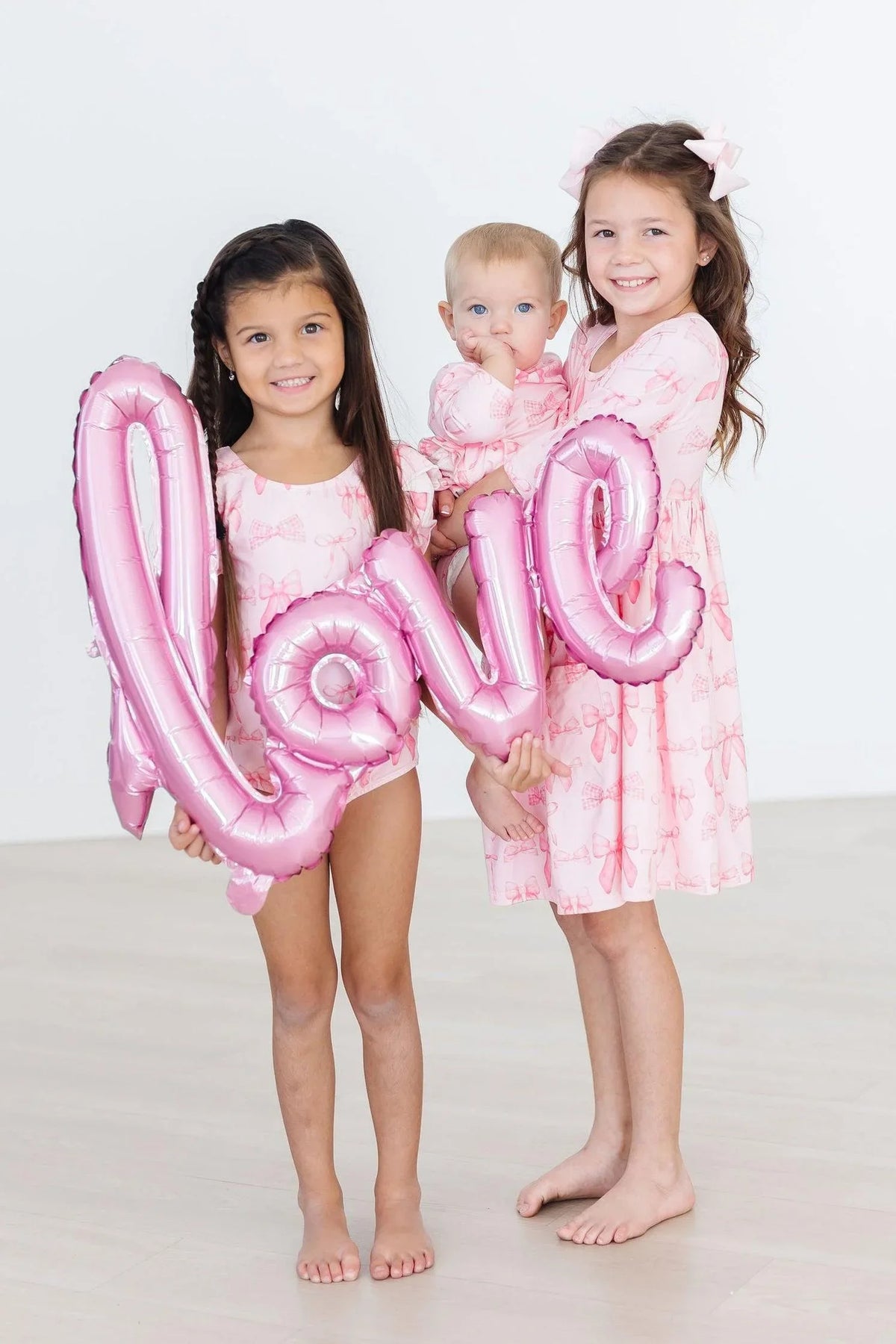 Two girls and a baby wearing pink baby girl bow dress and twirl bodysuit holding pink love balloon