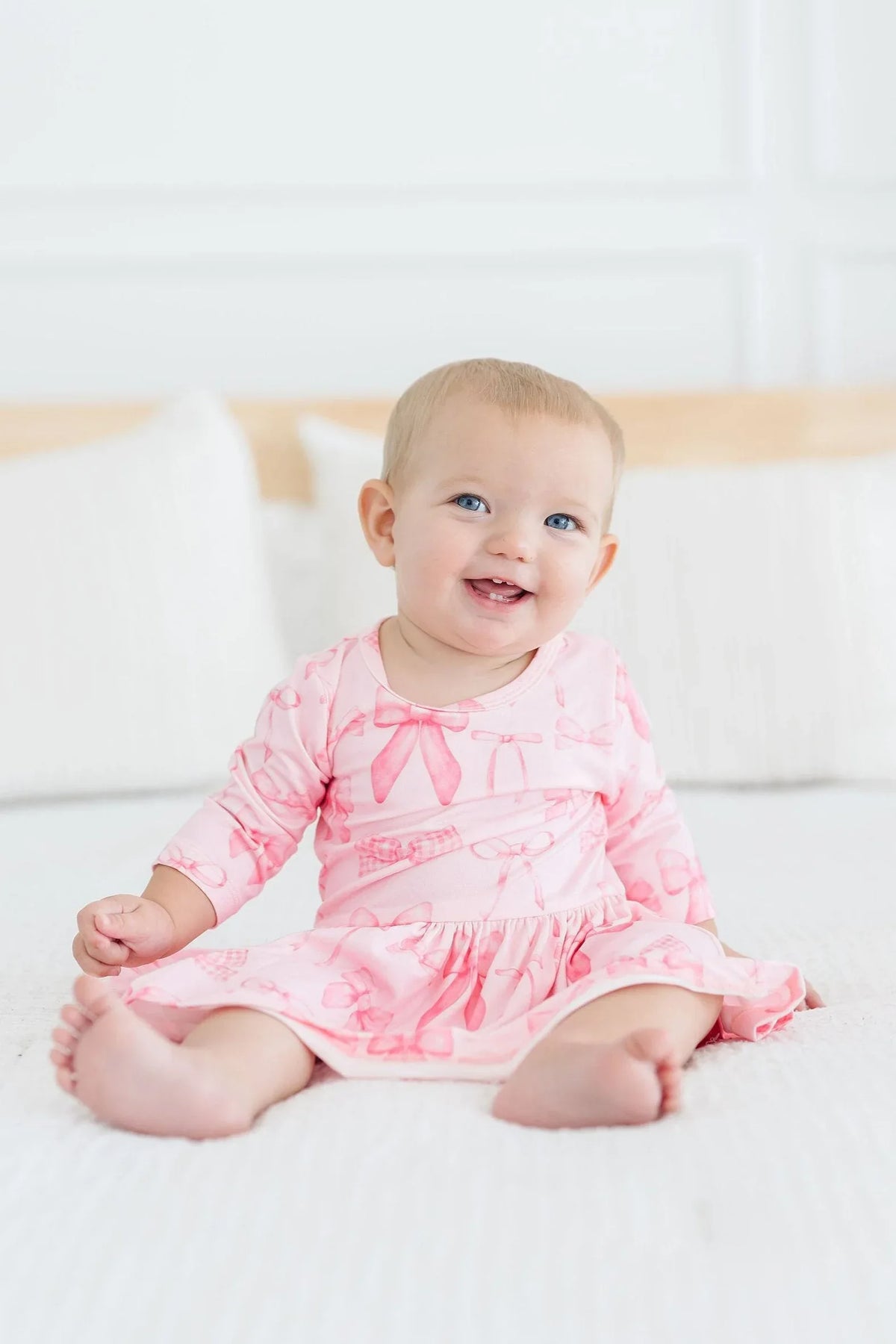Baby girl wearing pink Mila & Rose twirl bodysuit 3/4 sleeve sitting and smiling on white bed baby girl bow dress