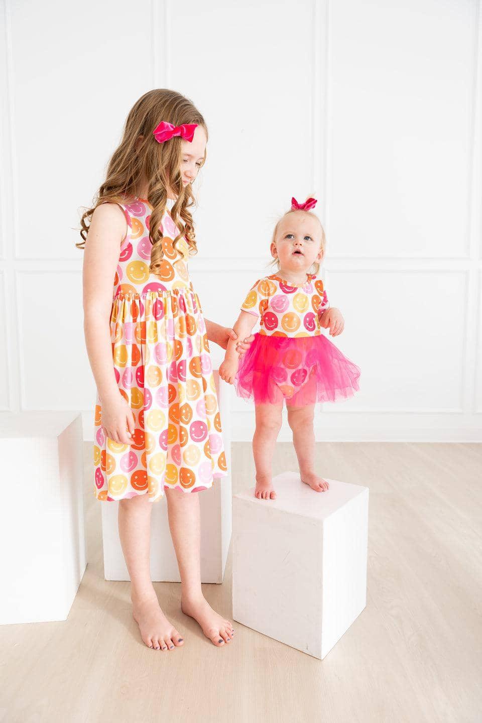 Baby girl wearing pink baby girl tutu bodysuit standing on white block holding older girl’s hand