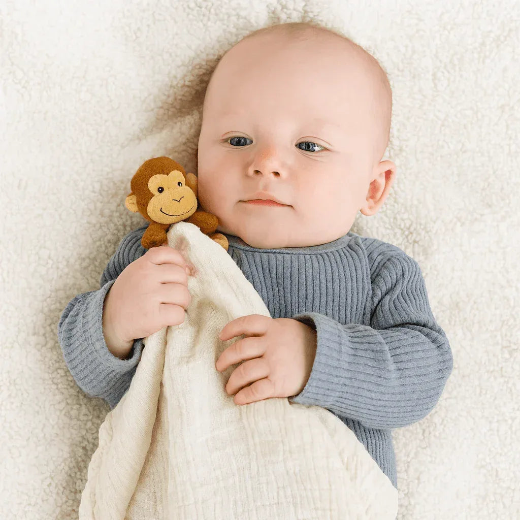 Baby holding cream cotton Me Lovey security blanket with brown monkey plush on soft white surface